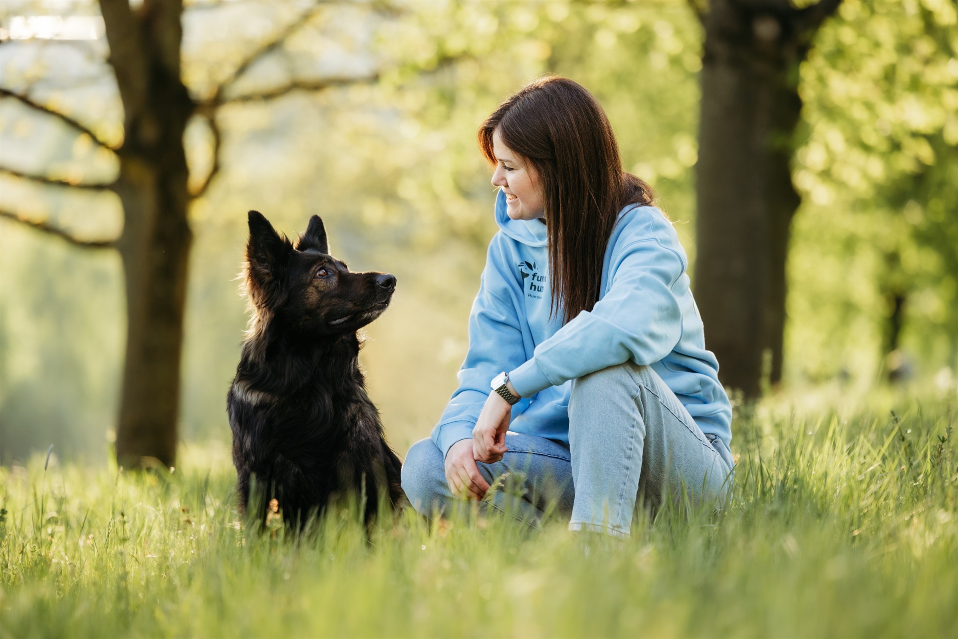 Frau mit hellblauen Hoody kniet in einer grünen Wiese. Ein schwarzer Hund sitzt ihr gegenüber. Sie blicken sich freundlich in die Augen.