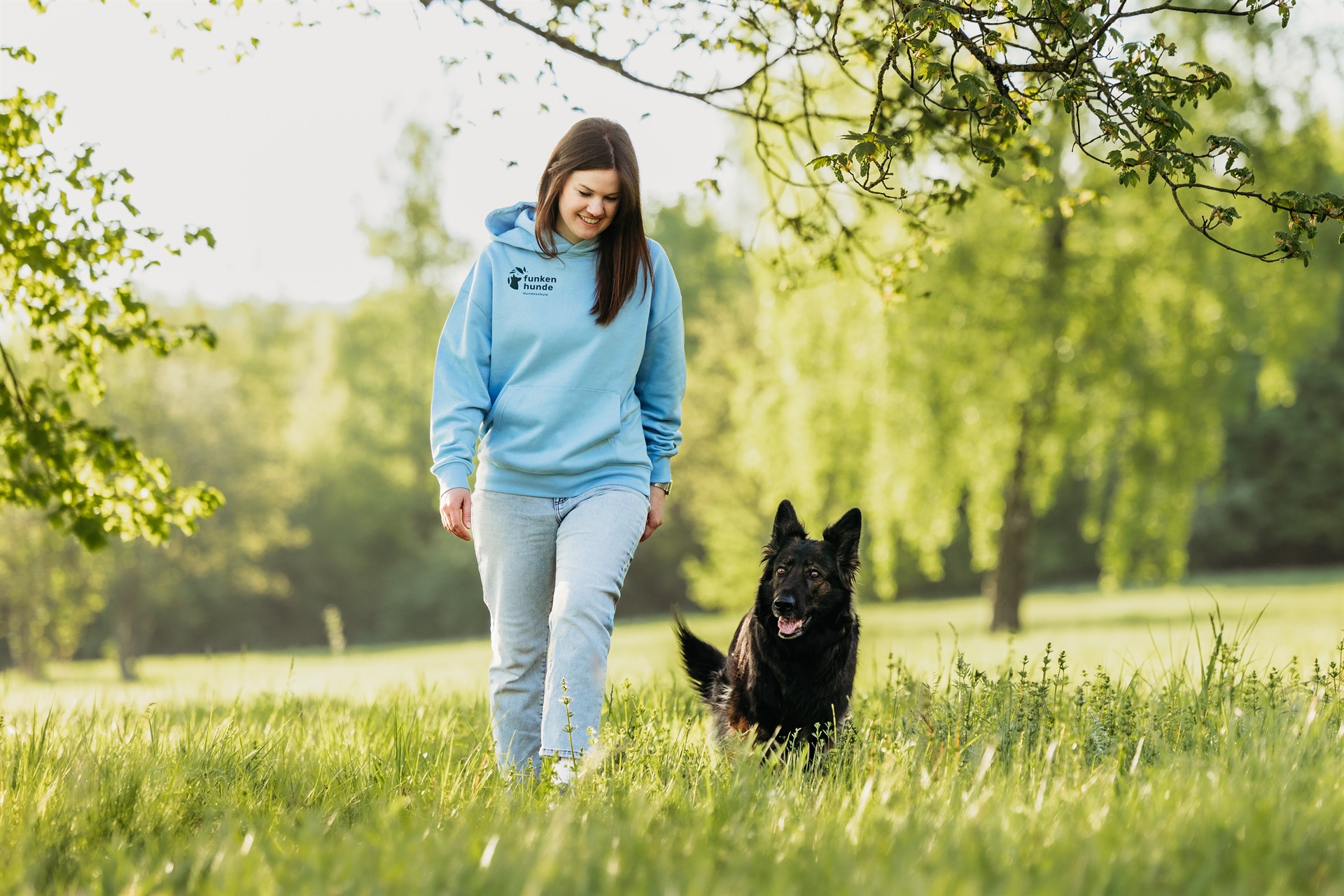 Frau in hellblauem Hoodie spaziert mit einem schwarzen Hund über eine sonnige Wiese unter blühenden Bäumen.