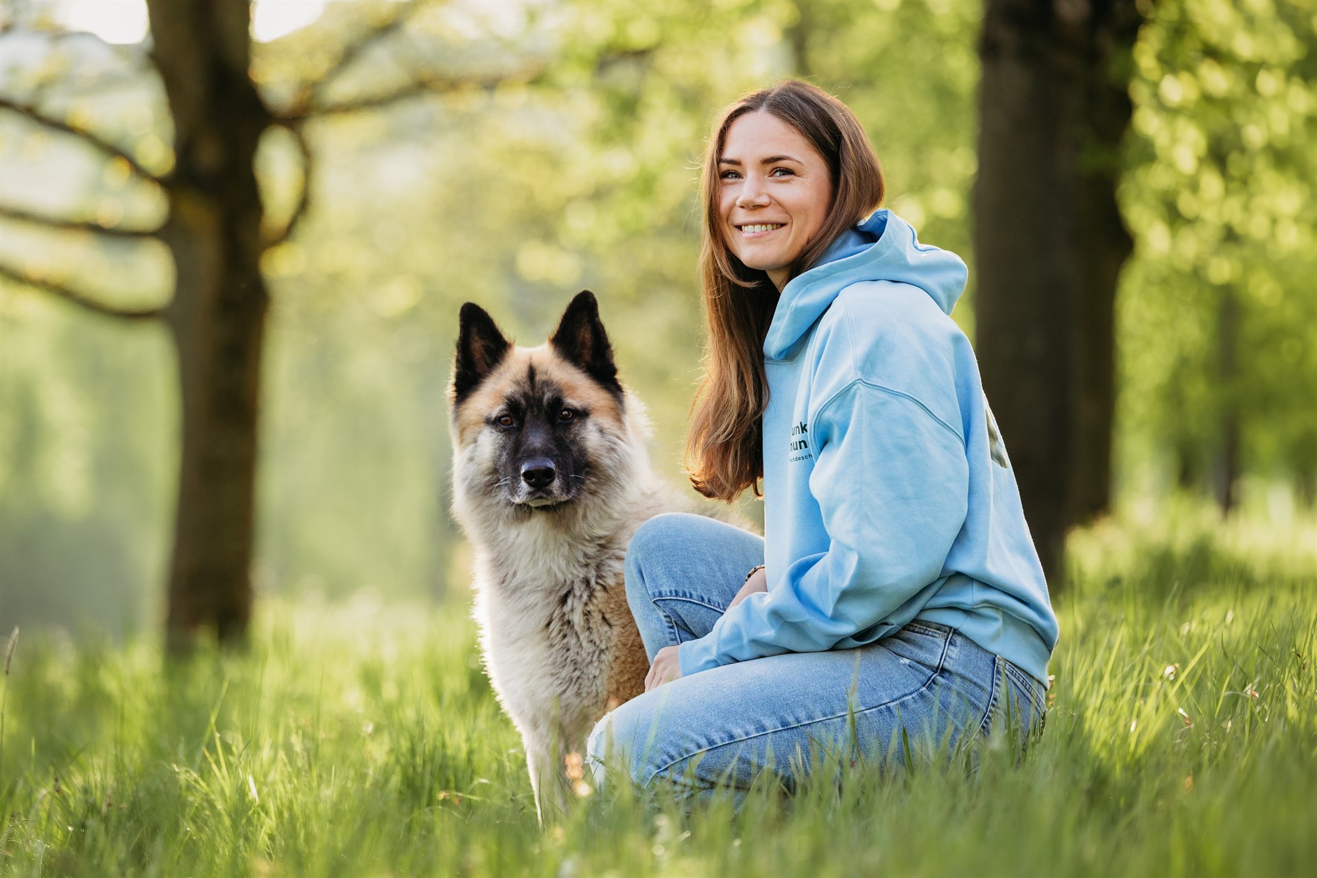 Frau mit hellblauen Hoody kniet in einer grünen Wiese. Ein weißer Hund sitzt neben ihr.
