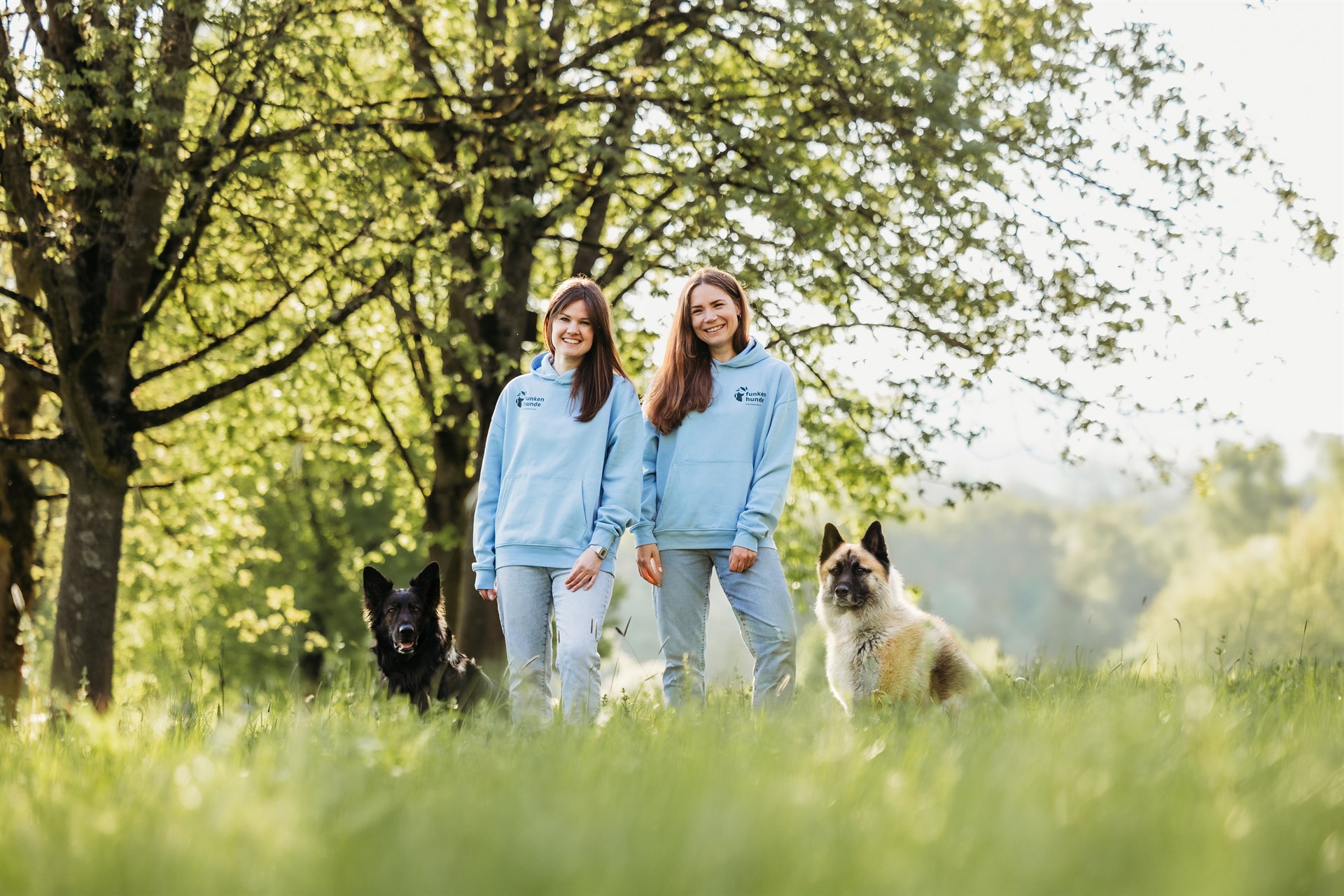 Zwei Frauen stehen auf einer Wiese. Links sitzt ein schwarzer Hund, rechts sitzt ein weißer Hund. Im Hintergrund stehen große Bäume.