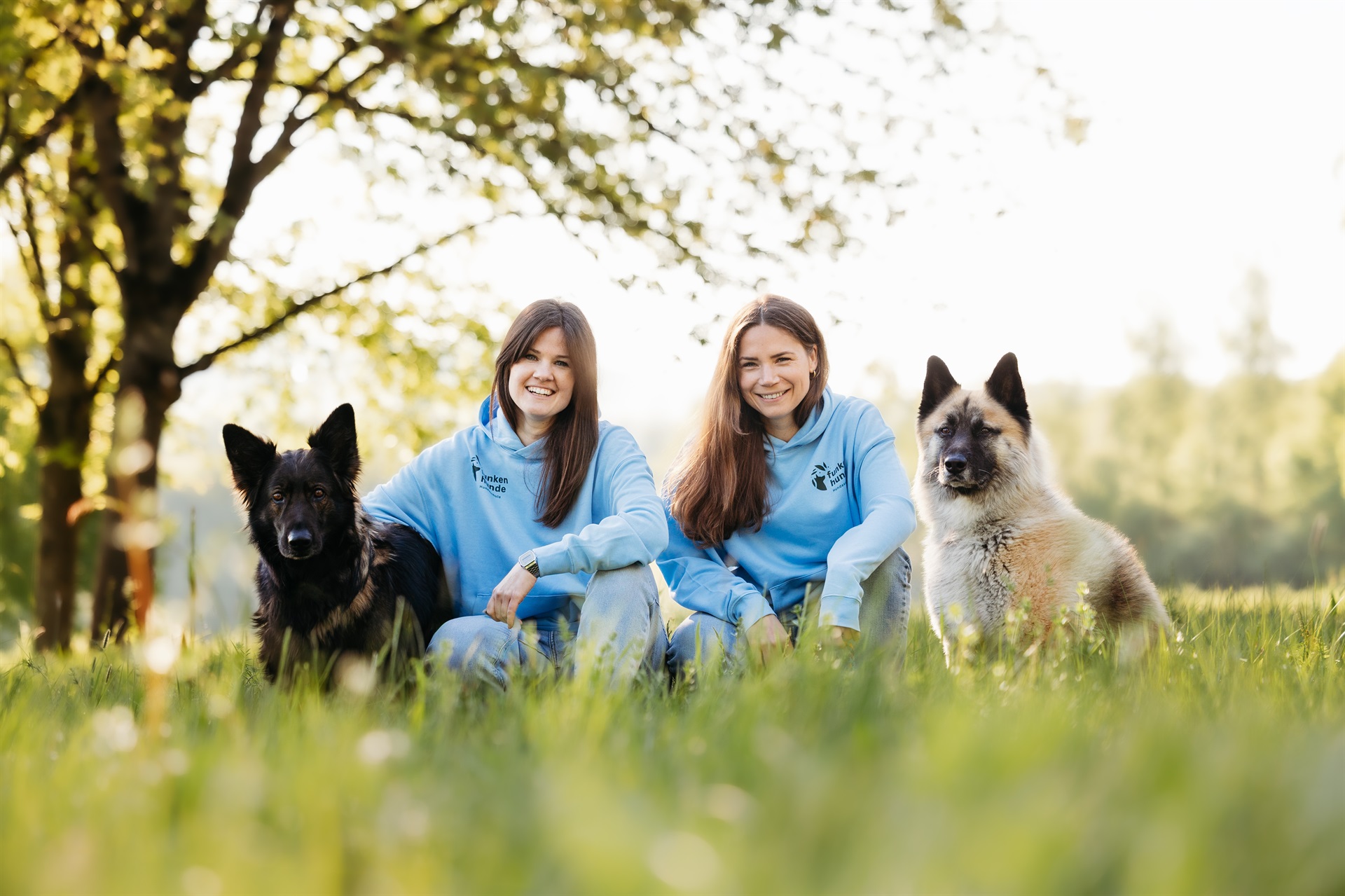 Zwei Frauen sitzen in einer Wiese. Links sitzt ein schwarzer Hund, rechts sitzt ein weißer Hund.