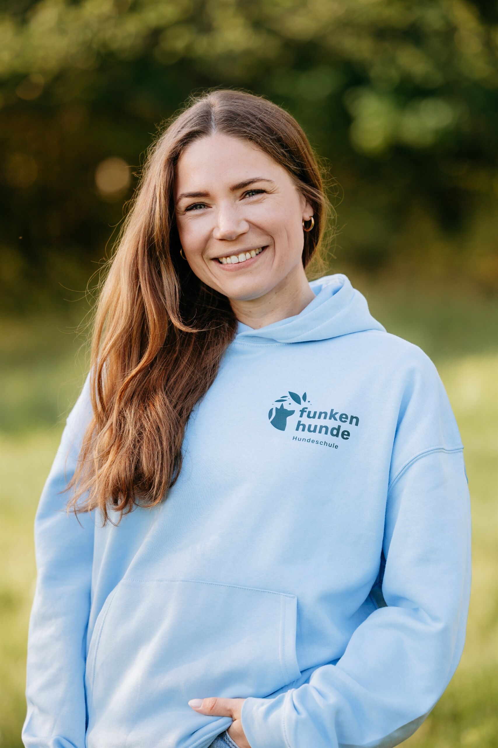 Frau mit hellblauem Hoody steht im grünen und lächelt in die Kamera.