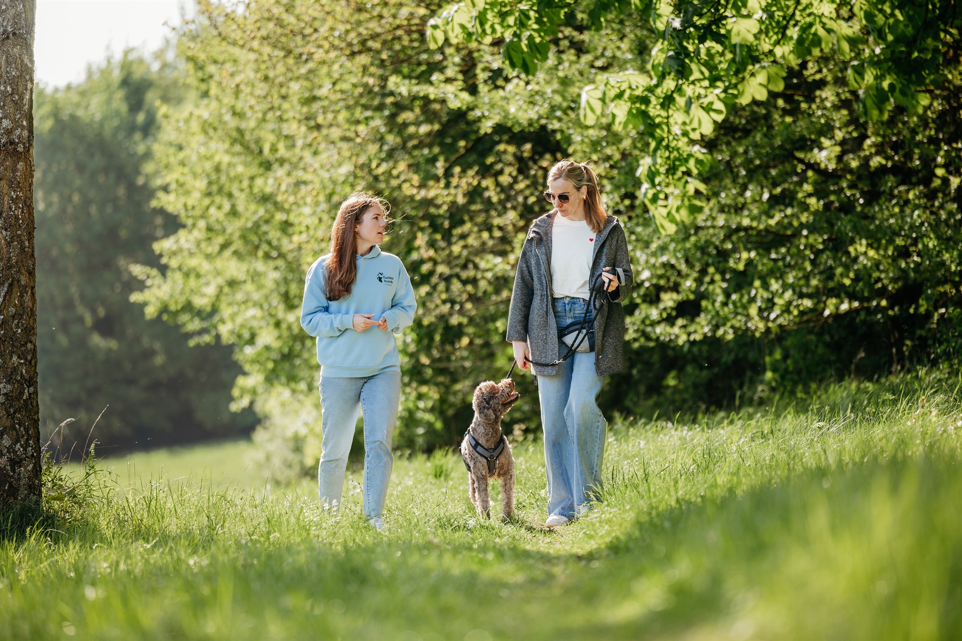 Zwei Frauen laufen auf einer hellgrünen Wiese. Eine Frau läuft mit einem angeleinten hellbraunen Hund. Im Hintergrund stehen Bäume.