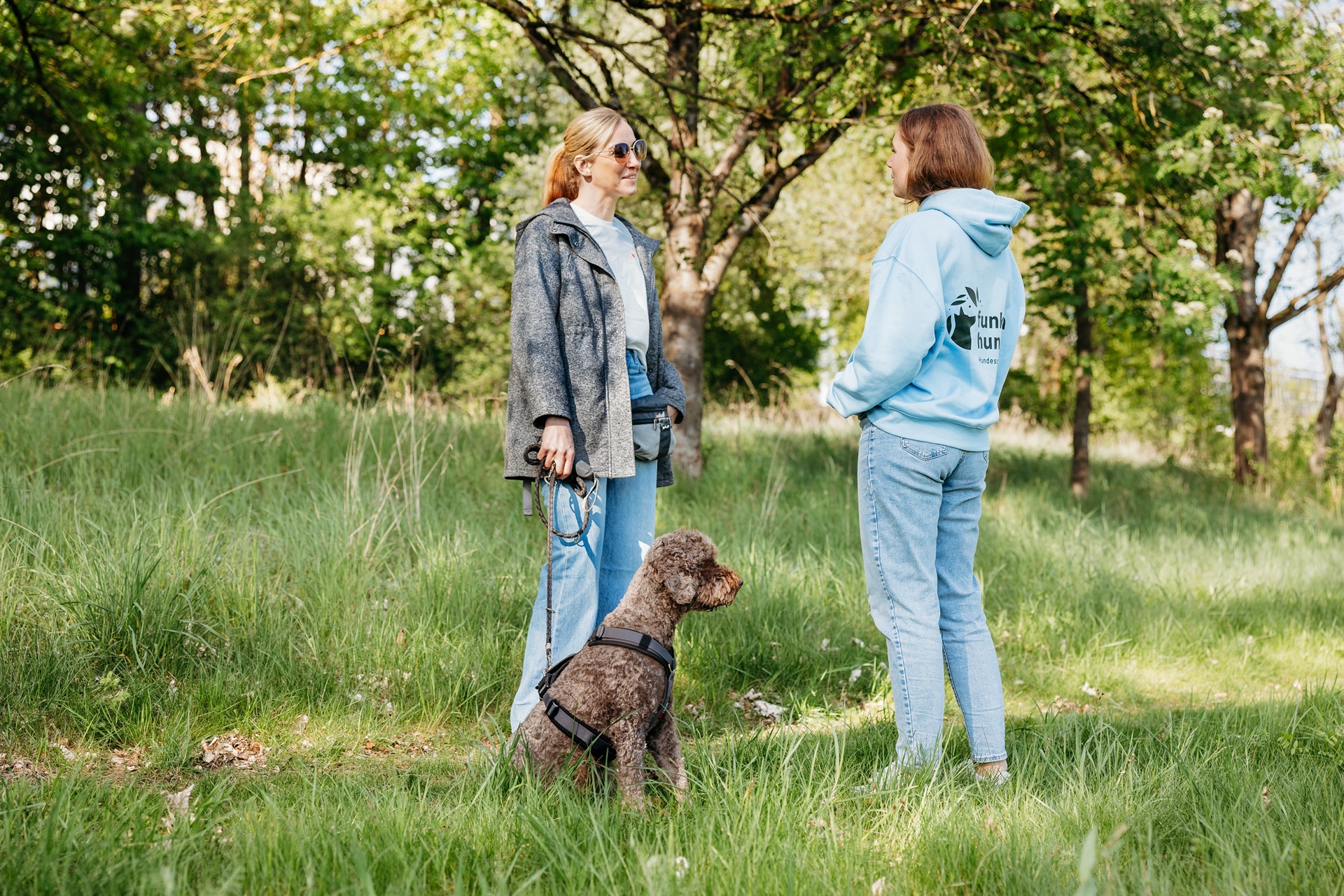Zwei Frauen unterhalten sich auf einer grünen Wiese umgeben von Bäumen. Eine Frau hält einen braunen Hund an der Leine. Der Hund blickt entspannt in die Ferne.